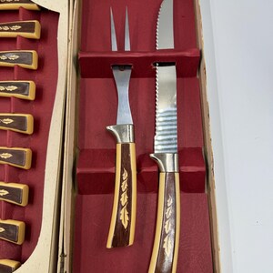 May include: Vintage carving set in a box. The set includes a carving fork, a serrated knife, and a carving knife. The handles are a light tan color with a dark brown wood inlay. The box is beige with a red interior.
