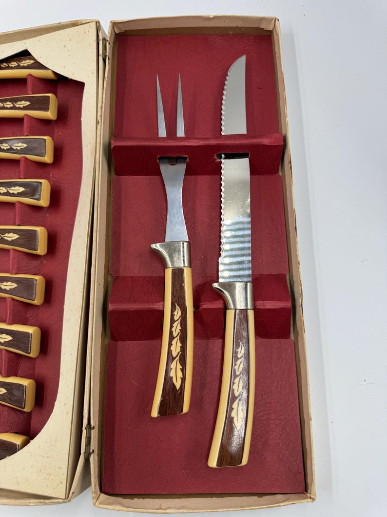 May include: Vintage carving set in a box. The set includes a carving fork, a serrated knife, and a carving knife. The handles are a light tan color with a dark brown wood inlay. The box is beige with a red interior.