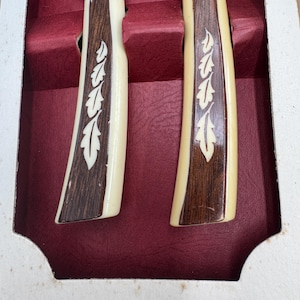 May include: Two knives with cream-colored handles, dark brown wood accents, and a silver blade. The handles feature a decorative leaf design. The knives are presented in a red velvet-lined box, suggesting a set for dining or display.
