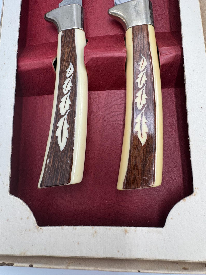 May include: Two knives with cream-colored handles, dark brown wood accents, and a silver blade. The handles feature a decorative leaf design. The knives are presented in a red velvet-lined box, suggesting a set for dining or display.