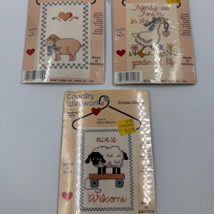 May include: Three Country Wireworks cross-stitch kits. One features a pig with a heart, another a goose with a heart, and the third has two sheep on a cart with the word "Welcome". Each kit is packaged in plastic.