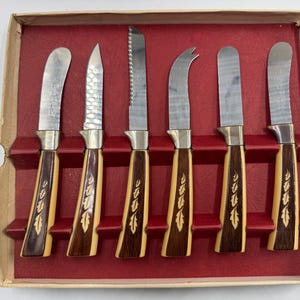 May include: A set of six vintage cheese knives in a cardboard box with a red velvet-like interior. The knives have stainless steel blades and handles with a light wood and dark wood grain design with a leaf pattern.