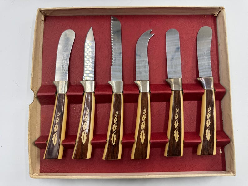 May include: A set of six vintage cheese knives in a cardboard box with a red velvet-like interior. The knives have stainless steel blades and handles with a light wood and dark wood grain design with a leaf pattern.
