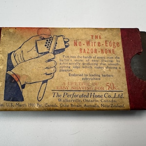 Puede incluir: Caja vintage "No-Wire-Edge Razor-Hone" con una ilustración de manos usando la piedra de afilar. El texto incluye "A Lifetime of Easy Shaving for 50c" y "The Perforated Hone Co., Ltd." La caja es marrón y roja.
