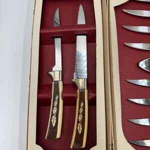 May include: A vintage set of steak knives in a cream-colored box with a red interior. The knives have stainless steel blades and handles with a cream and brown design. One knife has a serrated edge.