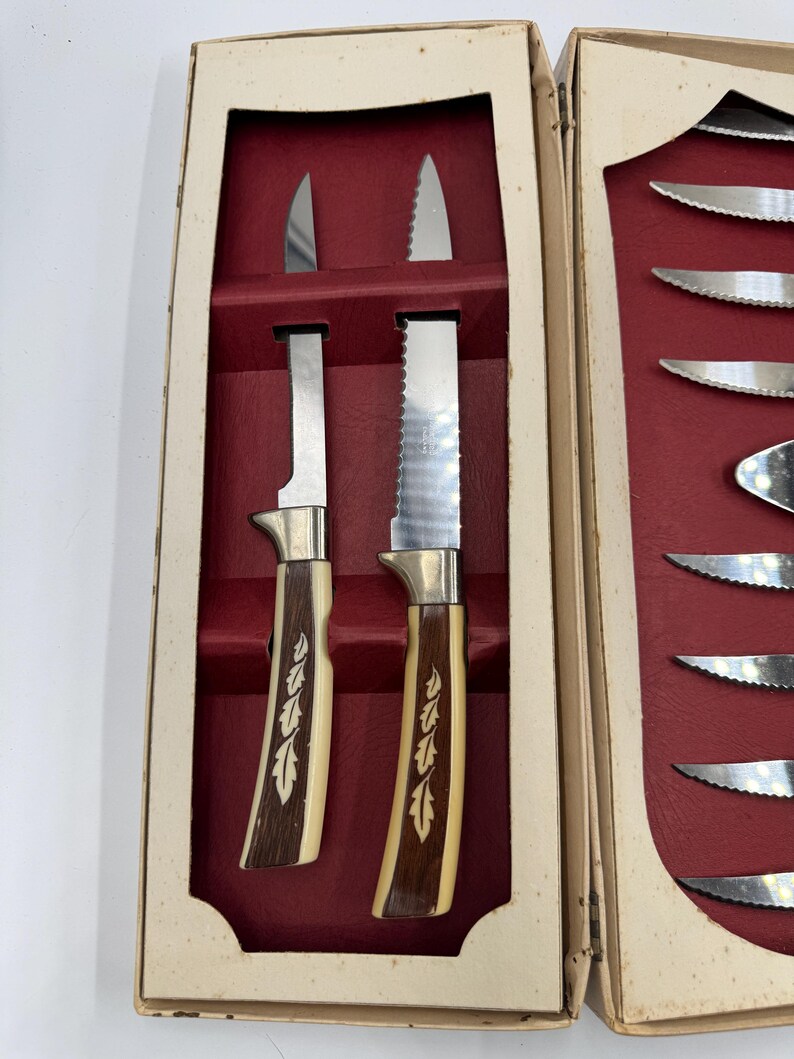 May include: A vintage set of steak knives in a cream-colored box with a red interior. The knives have stainless steel blades and handles with a cream and brown design. One knife has a serrated edge.