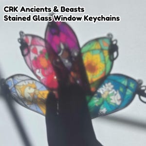Puede incluir: Una colección de cinco llaveros translúcidos con diseños de vidrieras, ilustraciones coloridas y contornos negros. El texto CRK Ancients & Beasts Stained Glass Window Keychains se muestra en la parte superior de la imagen.