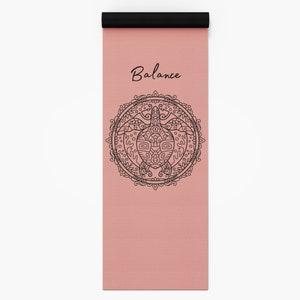 Puede incluir: Una esterilla de yoga rosa con un diseño de mandala negro que representa una tortuga. El texto "Balance" está impreso en negro en la parte superior de la esterilla.