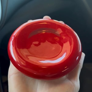 May include: A glossy, bright red, circular object with a slightly concave center. The object is held in a hand, and the background is a blurred interior, possibly a car. The object's surface reflects light.