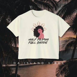 May include: White t-shirt featuring a graphic of a woman with brown hair and a pink top, with a sunburst design behind her. The text "HALF FILIPINA FULL BADDIE" is printed below the graphic. The text "DA ISLAND VIBES" is printed below the graphic.