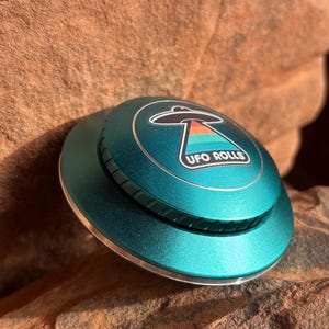 Peut inclure: Un moulin à herbe UFO Rolls en métal de couleur turquoise avec un bord argenté et un autocollant représentant un OVNI avec un motif arc-en-ciel et le texte "UFO ROLLS".
