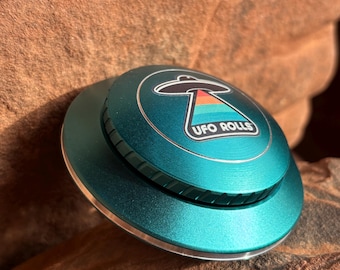 May include: A teal-colored metal UFO Rolls grinder with a silver rim and a sticker of a UFO with a rainbow design and the text "UFO ROLLS".