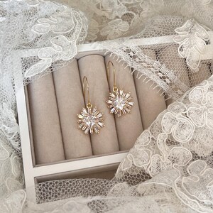 May include: A pair of gold dangle earrings with a clear crystal starburst design. The earrings are displayed in a white box with beige velvet lining.