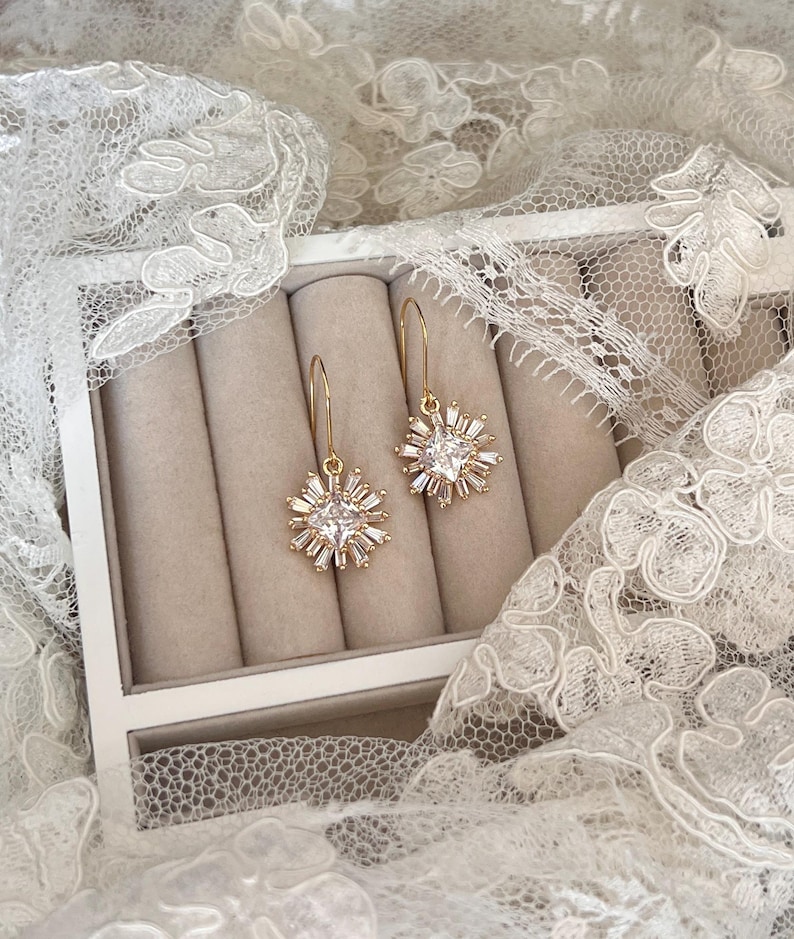 May include: A pair of gold dangle earrings with a clear crystal starburst design. The earrings are displayed in a white box with beige velvet lining.
