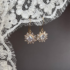 May include: A pair of gold dangle earrings with a clear square crystal surrounded by smaller crystals. The earrings are hanging from a white lace background.