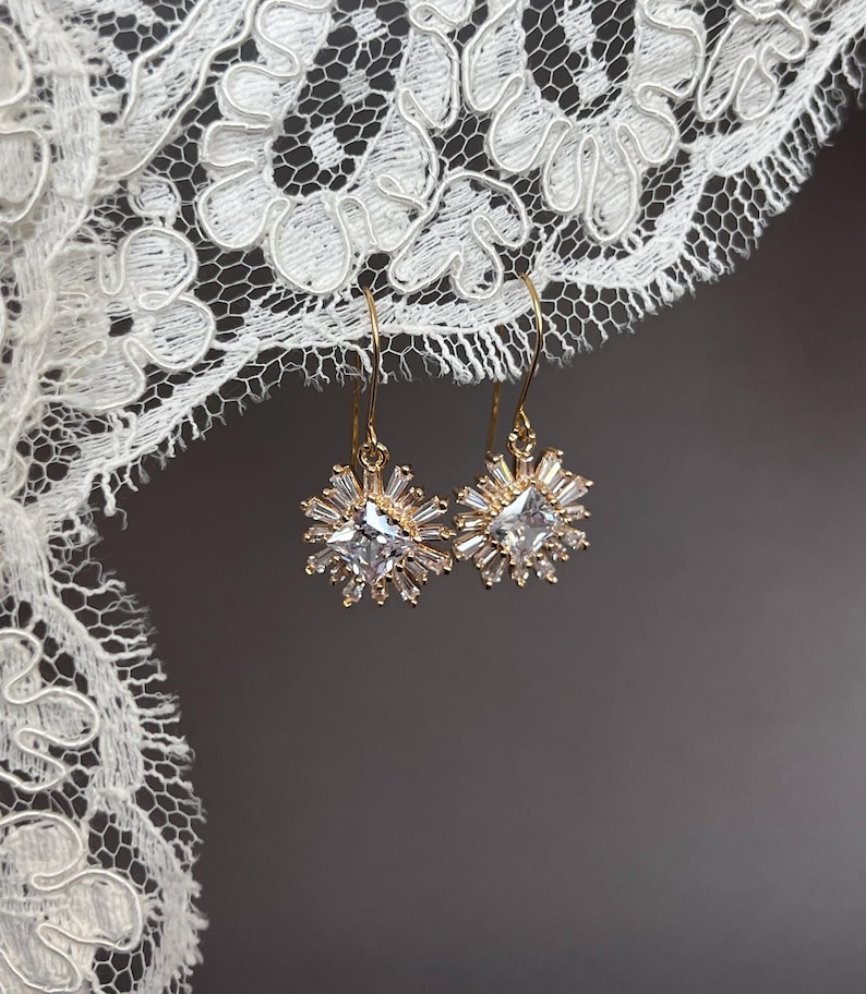 May include: A pair of gold dangle earrings with a clear square crystal surrounded by smaller crystals. The earrings are hanging from a white lace background.