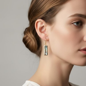 May include: Gold-tone rectangular earrings with a rectangular abalone shell inlay. The earrings have a simple hook closure. The abalone shell has iridescent blue, green, and silver tones. The earrings are shown on a person.