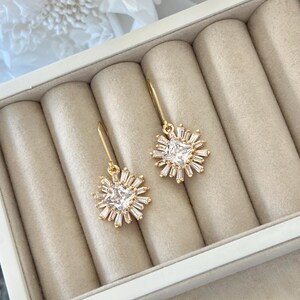 May include: A pair of gold earrings with a square-shaped clear crystal surrounded by smaller crystals in a starburst pattern.