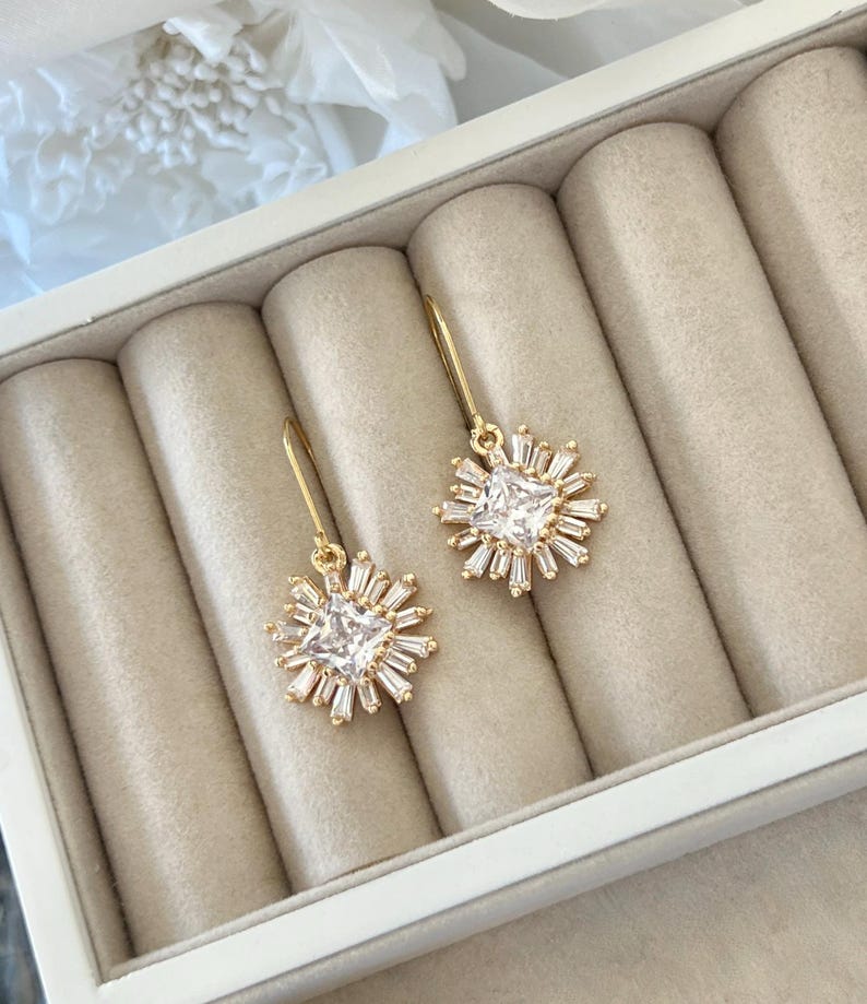 May include: A pair of gold earrings with a square-shaped clear crystal surrounded by smaller crystals in a starburst pattern.