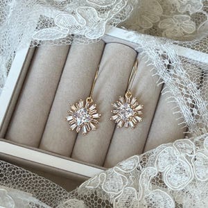 May include: A pair of gold earrings with a clear crystal starburst design. The earrings are in a white box with a velvet lining.