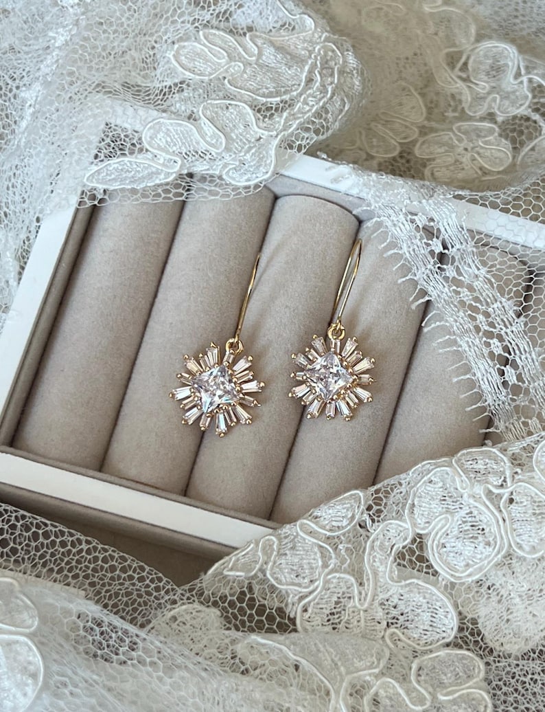 May include: A pair of gold earrings with a clear crystal starburst design. The earrings are in a white box with a velvet lining.