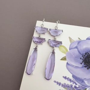 May include: A pair of silver dangle earrings with three tiers of purple faceted stones. The top two tiers are crescent shaped and the bottom tier is teardrop shaped.
