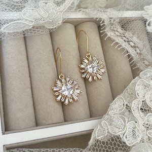 May include: A pair of gold dangle earrings with a square-shaped clear crystal surrounded by smaller crystals in a starburst design.