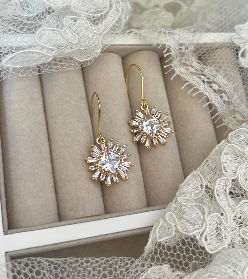 May include: A pair of gold dangle earrings with a square-shaped clear crystal surrounded by smaller crystals in a starburst design.