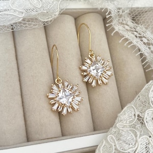 May include: A pair of gold dangle earrings with a square-shaped clear crystal surrounded by smaller crystals in a starburst design.