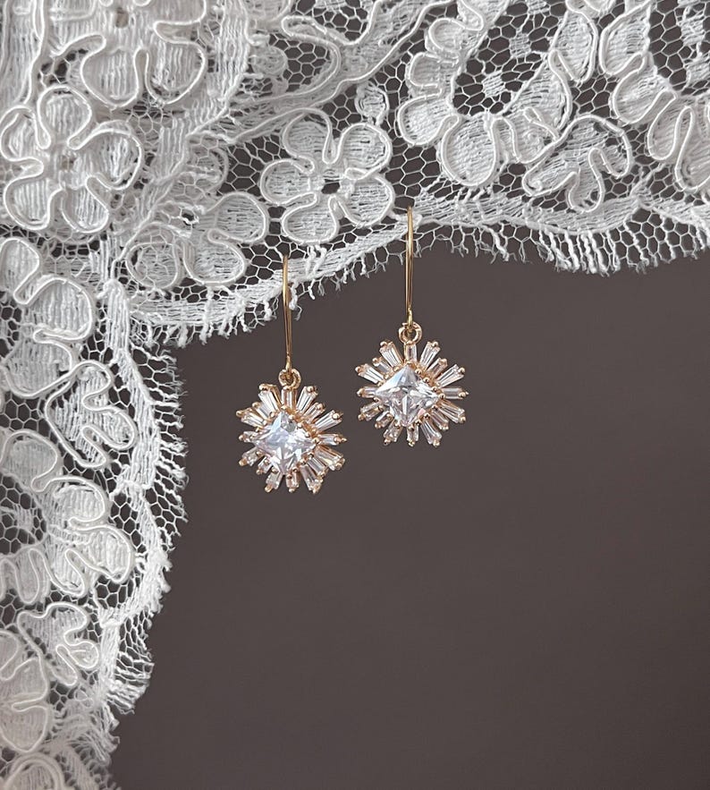May include: A pair of gold earrings with a square-shaped clear crystal surrounded by smaller crystals in a starburst design.