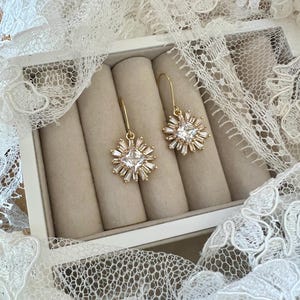 May include: A pair of gold earrings with a square-shaped crystal surrounded by smaller crystals in a starburst design. The earrings are displayed in a white box with beige velvet lining.