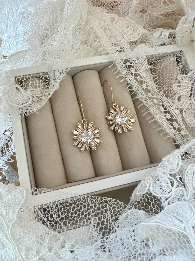 May include: A pair of gold earrings with a square-shaped crystal surrounded by smaller crystals in a starburst design. The earrings are displayed in a white box with beige velvet lining.
