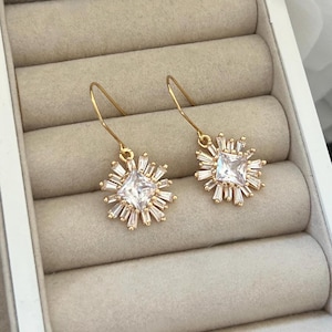 May include: A pair of gold earrings with a square-shaped clear crystal surrounded by smaller crystals in a starburst design.