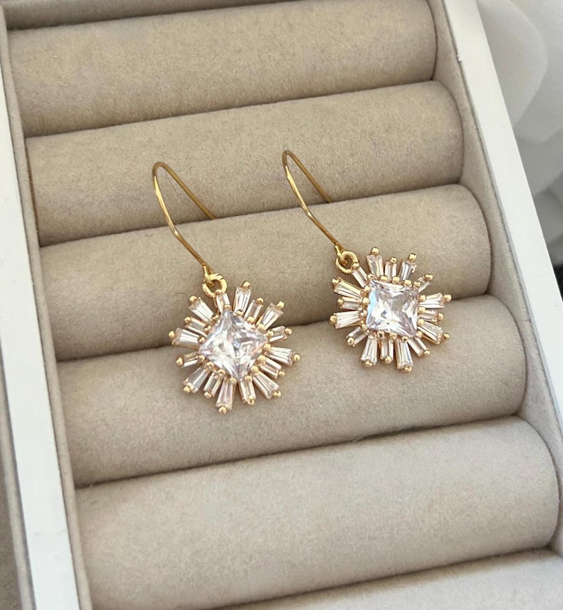 May include: A pair of gold earrings with a square-shaped clear crystal surrounded by smaller crystals in a starburst design.
