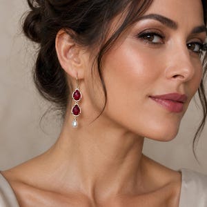 May include: A gold-toned drop earring featuring two stacked, faceted, deep red pear-shaped stones in metallic settings, finished with a single small white pearl dangling at the bottom, suspended from a simple gold-toned hook wire.