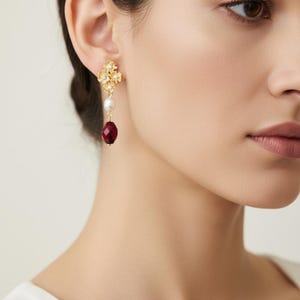 May include: Gold-toned drop earrings featuring a floral design with clear crystals. Each earring has a white pearl and a faceted, teardrop-shaped, deep red gemstone. The earrings are worn on a person's ear.