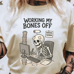 Może przedstawiać: Kremowa koszulka z napisem "WORKING MY BONES OFF" nad kreskówkowym szkieletem pracującym przy biurku. Szkielet otoczony jest filiżankami kawy, komputerem i karteczką "Deadline: Yesterday". Koszulka ma krótkie rękawy.