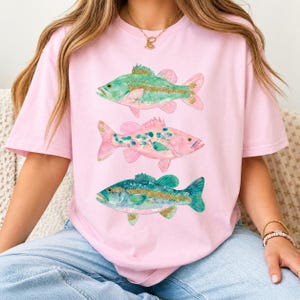 Puede incluir: Una camiseta de manga corta de color rosa claro con un estampado gráfico de tres peces apilados verticalmente. Los peces superior e inferior son de color verde azulado y verde con detalles dorados, mientras que el central es rosa.