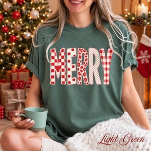 May include: A dark green short-sleeve t-shirt featuring the word MERRY printed in large block letters. Each letter displays a different red and white pattern, including checkered, polka dot, floral, and gingham designs on the fabric.