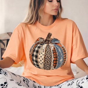 Könnte beinhalten: Ein pfirsichfarbenes T-Shirt mit kurzen Ärmeln und einem großen Kürbis-Motiv auf der Vorderseite. Der Kürbis besteht aus verschiedenen vertikalen Stoffmustern, darunter schwarz-weißes Karomuster, Leopardenmuster, Denim und Spitzenbesatz.