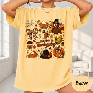 May include: A butter yellow short-sleeve t-shirt featuring a graphic print with the text Happy Thanksgiving. The design includes a turkey, pumpkin, pie, coffee cup, plaid bow, and autumn leaves in shades of orange, brown, and black.