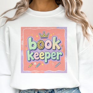 May include: White sweatshirt with a colourful graphic that reads "book keeper." The design includes a crown, pencils, and stars. The text is in a playful font with pastel colours on a pink and purple background.