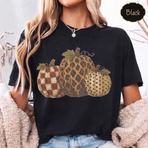 Peut inclure: Un t-shirt noir à manches courtes avec une impression graphique de trois citrouilles. La citrouille de gauche a un motif à carreaux marron et beige, celle du centre un imprimé léopard marron et celle de droite un motif à pois beige.