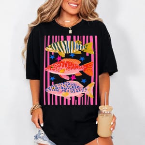 Puede incluir: Una camiseta negra de manga corta con un estampado gr&aacute;fico de tres peces estilizados apilados verticalmente. Los peces est&aacute;n representados en amarillo, naranja y rosa con patrones de leopardo y rayas, sobre un fondo de rayas verticales rosas y peque&ntilde;as estrellas azules y rosas.