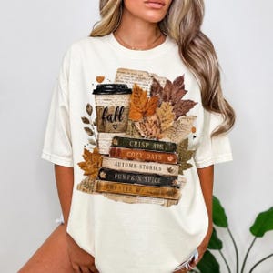May include: An off-white short-sleeve t-shirt featuring a graphic print of a coffee cup labeled fall with a heart, surrounded by autumn leaves and a stack of books titled Crisp Air, Cozy Days, Autumn Stories, Pumpkin Spice, and Sweater Weather.