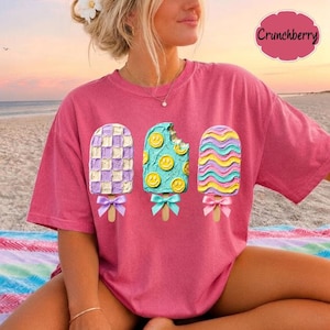 May include: A pink t-shirt featuring three popsicle graphics with textured frosting. The left popsicle has a purple and white checkerboard pattern, the center is teal with yellow smiley faces, and the right has wavy pink and blue stripes.