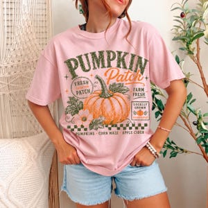 May include: A light pink short-sleeve t-shirt featuring a graphic of an orange pumpkin with green leaves and text that reads PUMPKIN Patch, FRESH FROM THE PATCH, FARM FRESH, LOCALLY GROWN, AUTUMN HARVEST, PUMPKINS, CORN MAZE, and APPLE CIDER.
