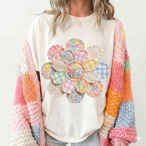 May include: A cream-coloured t-shirt with a large, colourful flower design made from various patterned fabric circles. A multi-coloured, chunky knit cardigan in shades of pink, orange, yellow, and blue is worn over the shirt. The flower design includes floral, gingham, and lace patterns.