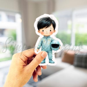 May include: A sticker featuring a cartoon illustration of a child in a light blue racing suit, holding a matching helmet. The child has black hair and a friendly expression. The sticker is white with a rounded edge.