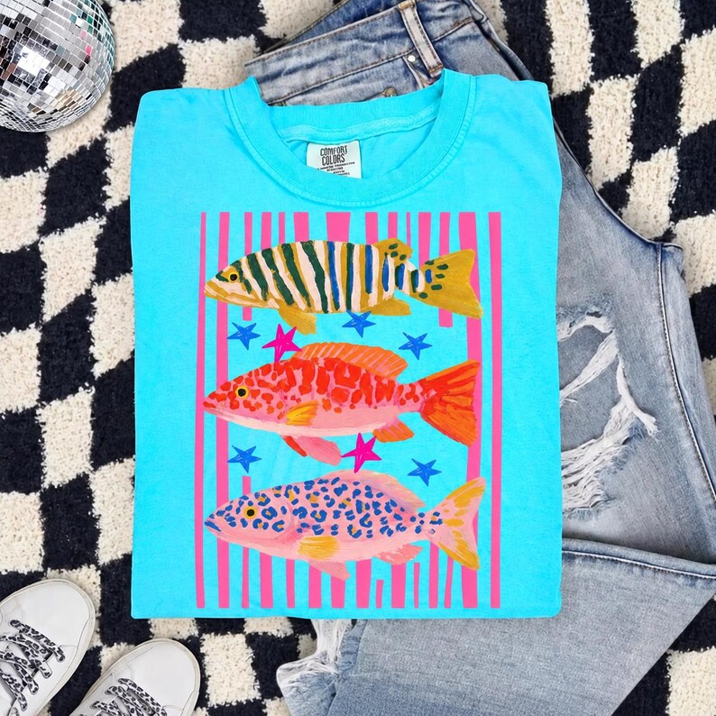 Puede incluir: Una camiseta de color turquesa brillante con un estampado gr&aacute;fico de tres peces estilizados apilados verticalmente. Los peces tienen patrones distintos en amarillo, naranja y rosa, sobre un fondo de rayas verticales rosas y peque&ntilde;as estrellas azules.